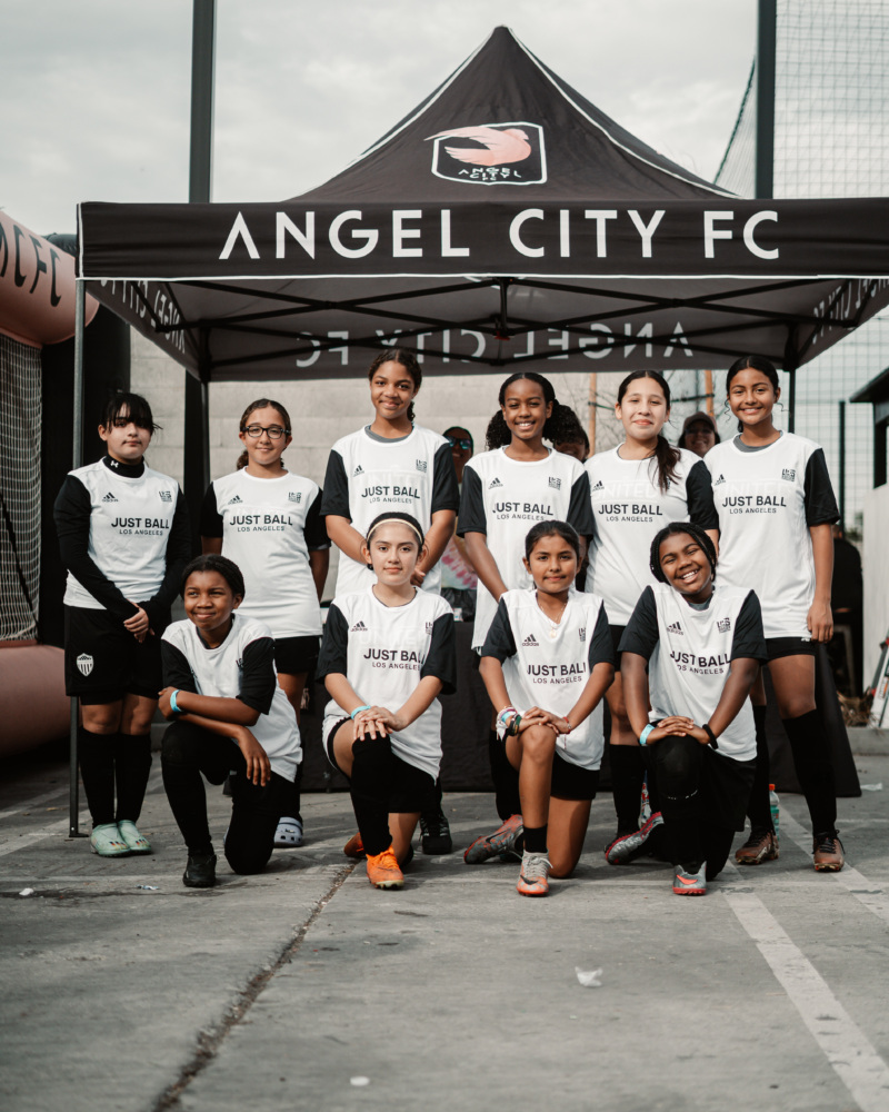 A group of ten girls in two rows smiles in front of an Angel City FC tent.